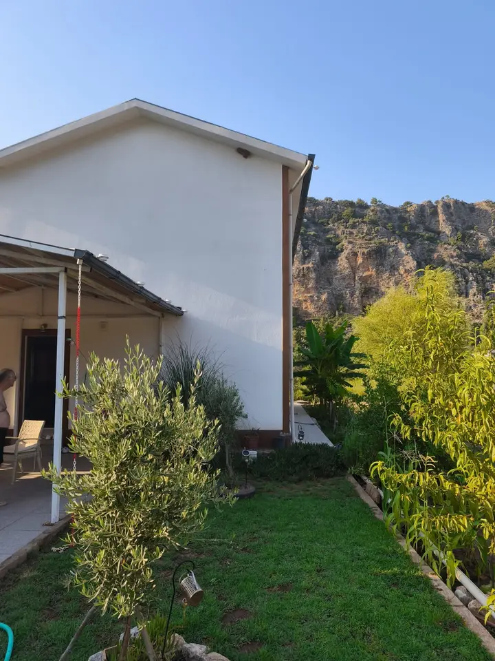 Maviev, a white two-storey house with a covered patio, a lawn and an olive tree, below the limestone cliffs