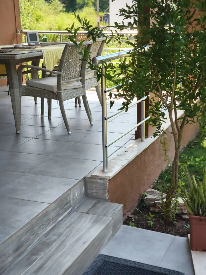 Tiled terrace with a table and wicker chairs, next to a young tree and the lawn